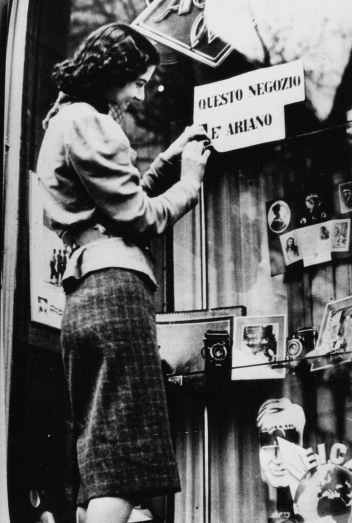 Het meisje hangt een papier op met de tekst: ‘Deze winkel is Arisch’. Rome, 1938.