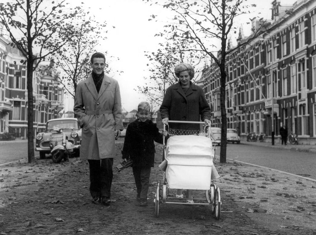 Wandelend gezin (vader, moeder, zoontje), met moeder achter de kinderwagen. Nederland, plaats onbekend, ca. 1960